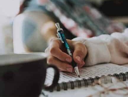 A person writes in a notebook while a cup of coffee sits nearby, indoors.