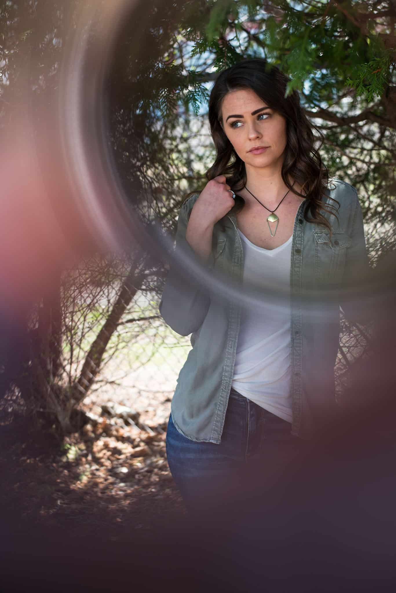 Young brunette woman in denim jacket and white shirt in front of chainlink fence and trees