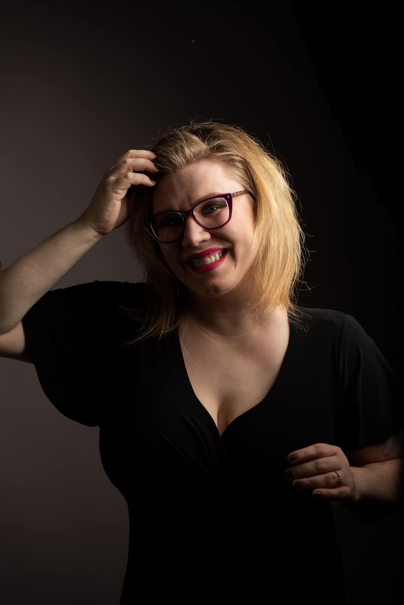 Blonde woman in black dress with purple glasses and pink lipstick touching hair and smiling