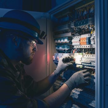 Male electrician working in switchboard with electrical connecting cable. Young adult electrical engineer in special clothes with flashlight on helmet in dark room with emergency lights in background.