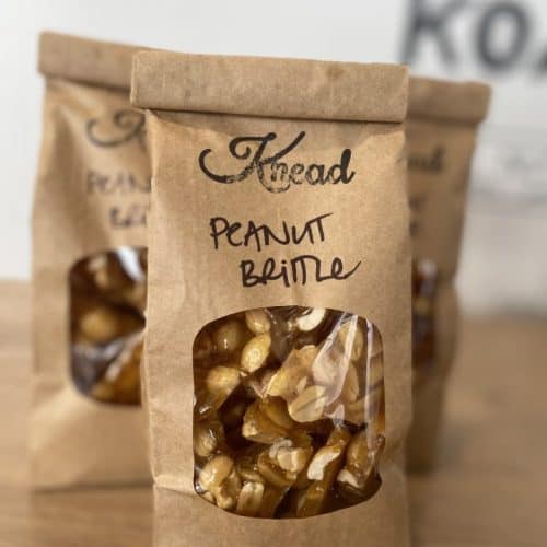 Close-up of handcrafted peanut brittle in a rustic paper bag with a transparent window, showcasing crunchy caramelized nuts.