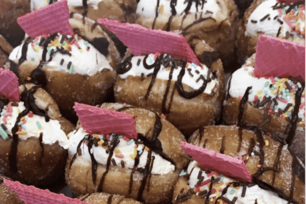 Close-up of sweet donuts topped with whipped cream, chocolate drizzle, colorful sprinkles, and pink wafers.