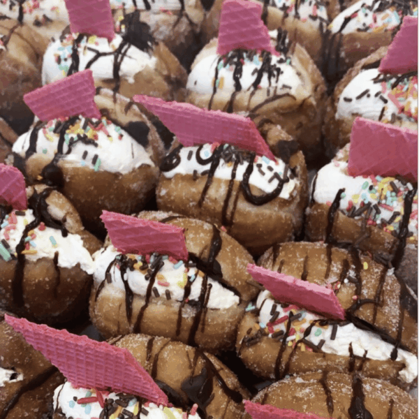 Close-up of sweet donuts topped with whipped cream, chocolate drizzle, colorful sprinkles, and pink wafers.