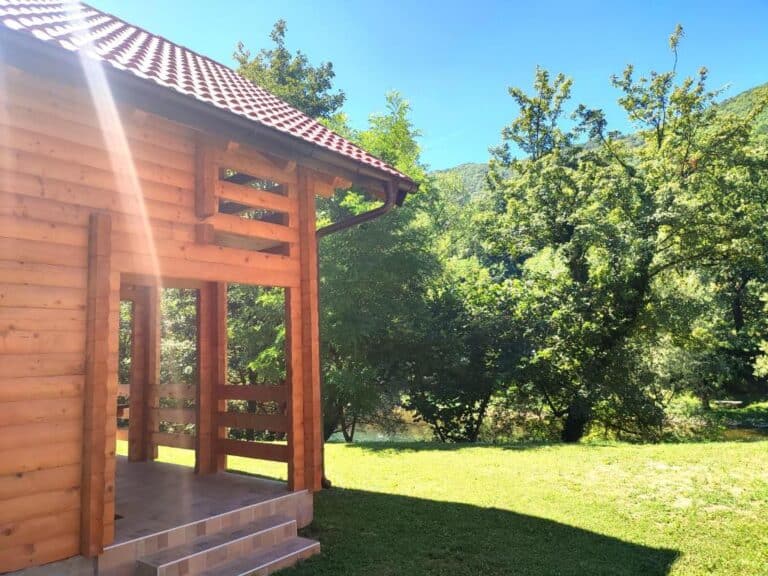 Drvena kuća Apartmana Kajkut u prirodnom okruženju uz rijeku Vrbas, okružena zelenilom i sunčanim ambijentom.