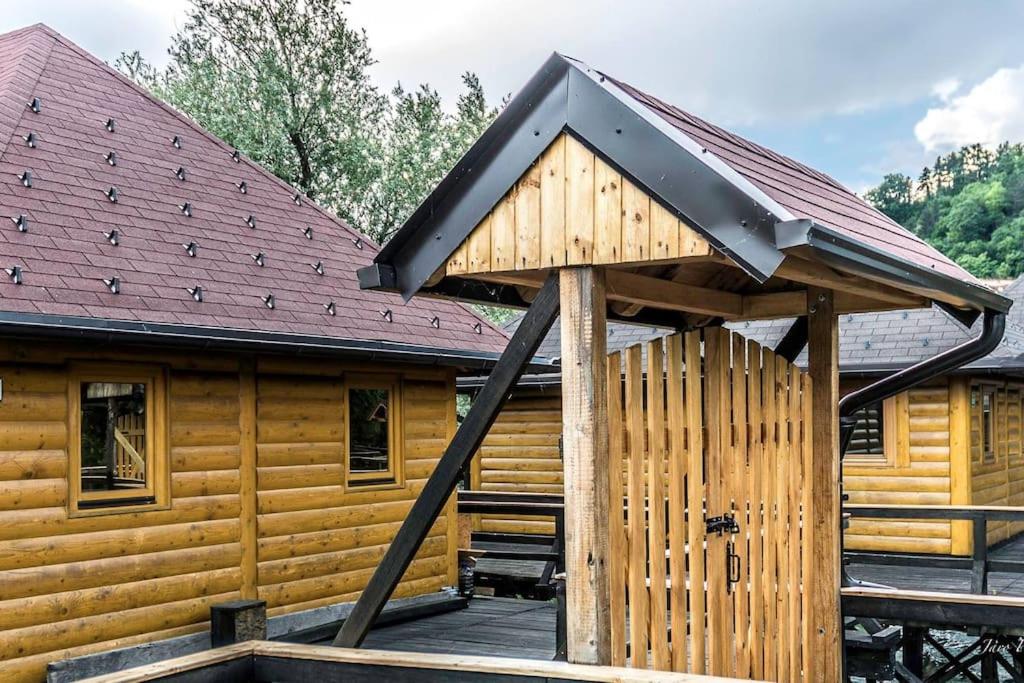 Drvene kućice s tradicionalnim krovovima i drvenom kapijom u prirodnom ambijentu, idealne za odmor uz rijeku.