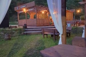 Vanjski pogled na AutoCamp Aqualand Bihać s rustikalnim drvenim stolovima i restoranom u prirodnom okruženju Nacionalnog parka Una.
