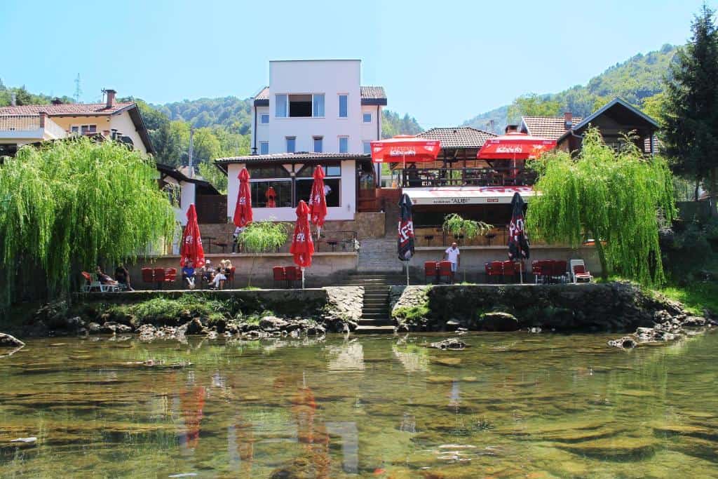 Motel Alibi u Banjoj Luci, smješten uz rijeku, s terasom, crvenim suncobranima i zelenim prirodnim okruženjem.