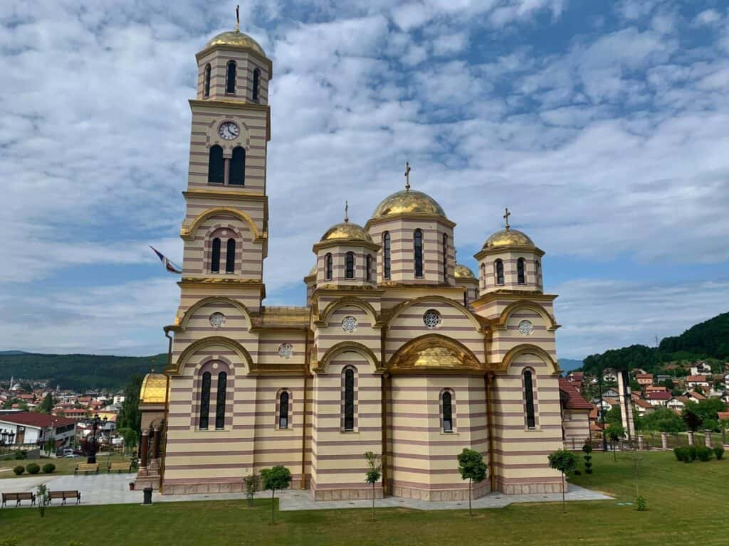 Crkva Svetog Save u Mrkonjić Gradu prepoznatljivim zlatnim kupolama, okruženim zelenim pejzažom i vedrim nebom.