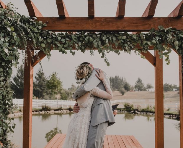A couple embraces under a wooden arbor decorated with greenery by a pond at Wind Mountain Ranch, creating a peaceful outdoor wedding scene.