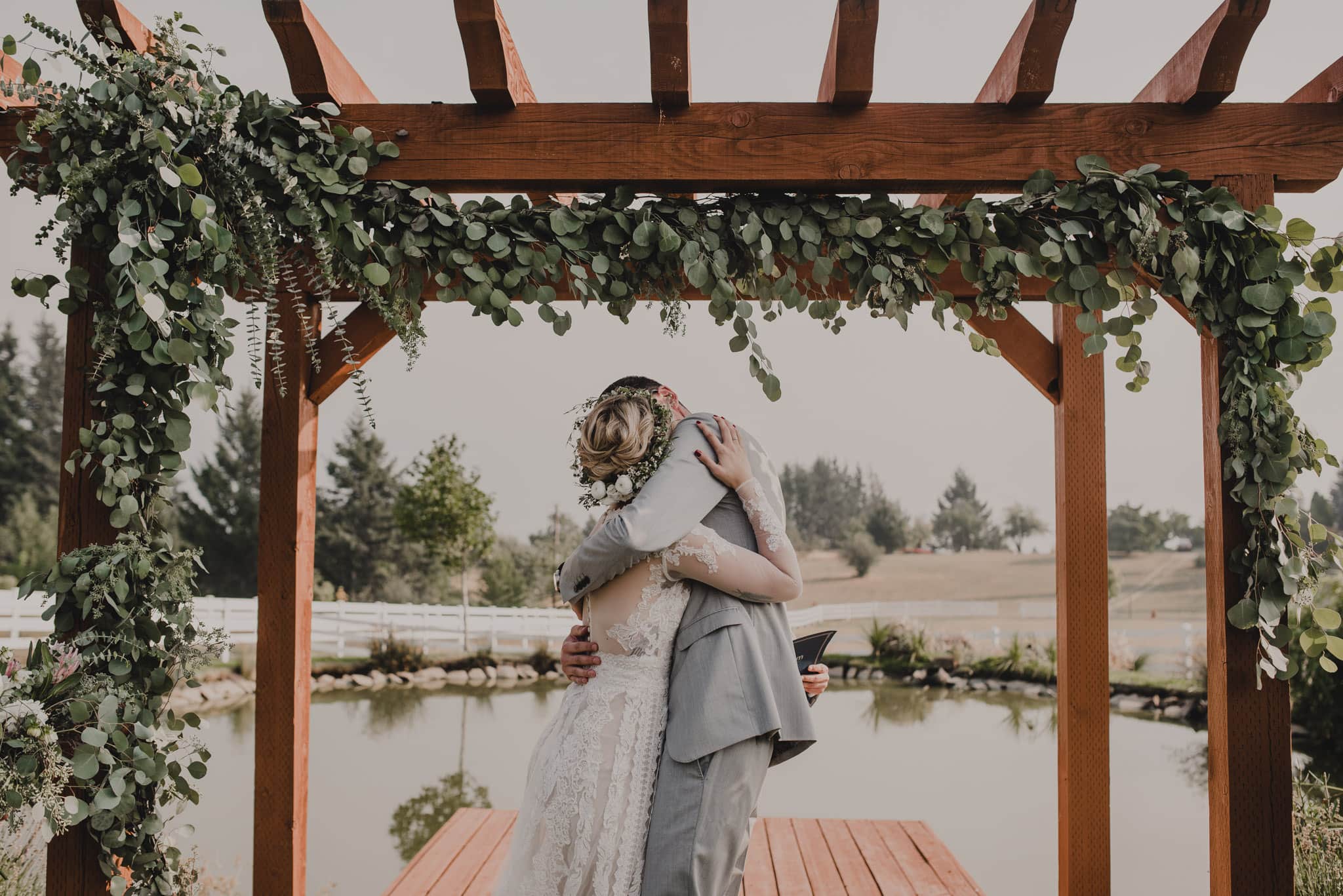 A couple embraces under a wooden arbor decorated with greenery by a pond at Wind Mountain Ranch, creating a peaceful outdoor wedding scene.