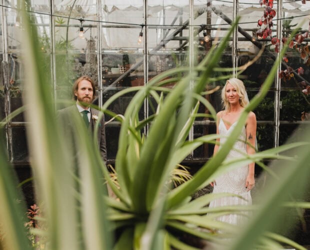 The bride and groom pose amidst the lush greenery of Pomarius Nursery, their portraits elegantly framed by large plant leaves, before heading to their wedding at Castaway Portland.