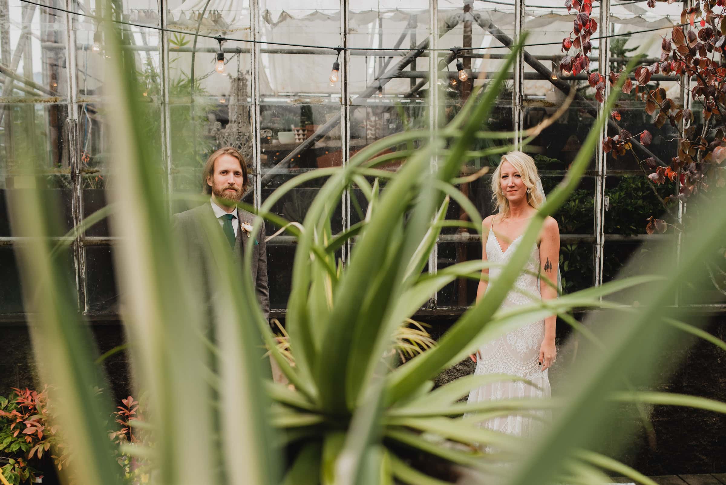 The bride and groom pose amidst the lush greenery of Pomarius Nursery, their portraits elegantly framed by large plant leaves, before heading to their wedding at Castaway Portland.