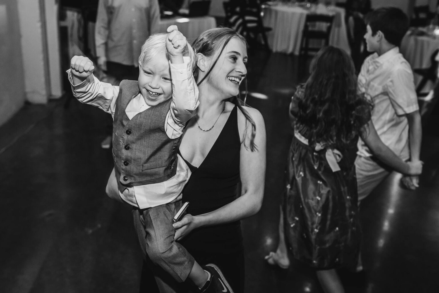 A woman joyfully holds a smiling child in her arms at a lively dance gathering, capturing a delightful moment akin to wedding photos at Castaway Portland.