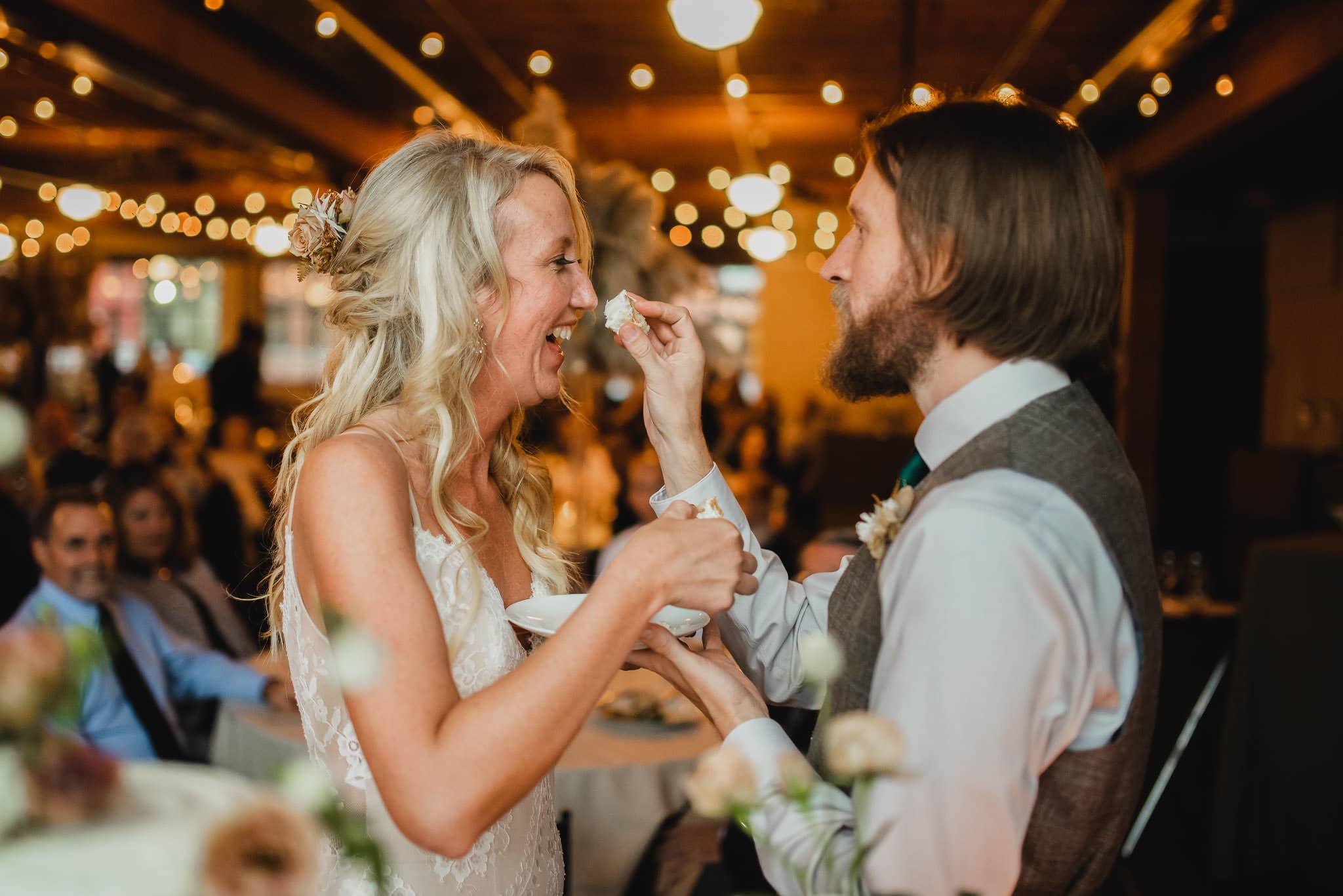 The bride and groom feed each other cake at their wedding reception, surrounded by decorative lights. Capturing this moment would make for stunning wedding photos at Castaway Portland.