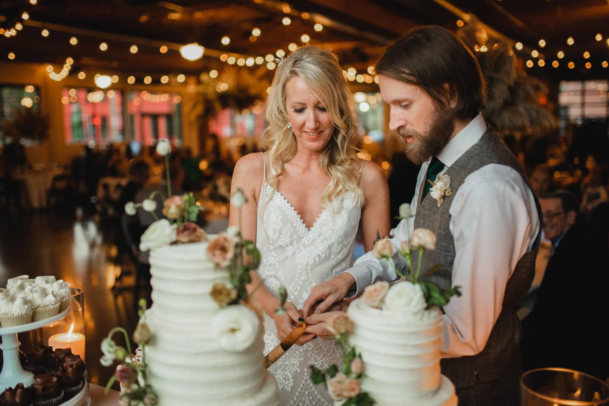 A couple cutting a tiered wedding cake adorned with flowers, surrounded by fairy lights in a warmly lit venue, captures the perfect moment for their wedding photos at Castaway Portland.