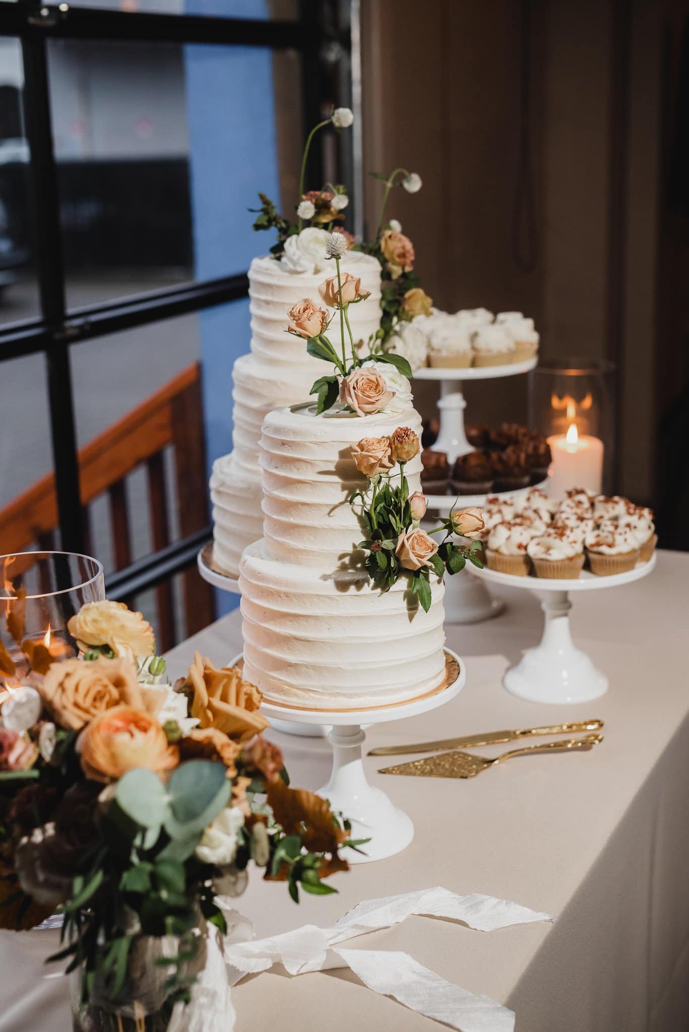A beautifully adorned wedding cake featuring flowers sits elegantly on a table, surrounded by glowing candles and adorable cupcakes, basking in natural light. Perfect for capturing stunning wedding photos at Castaway Portland.