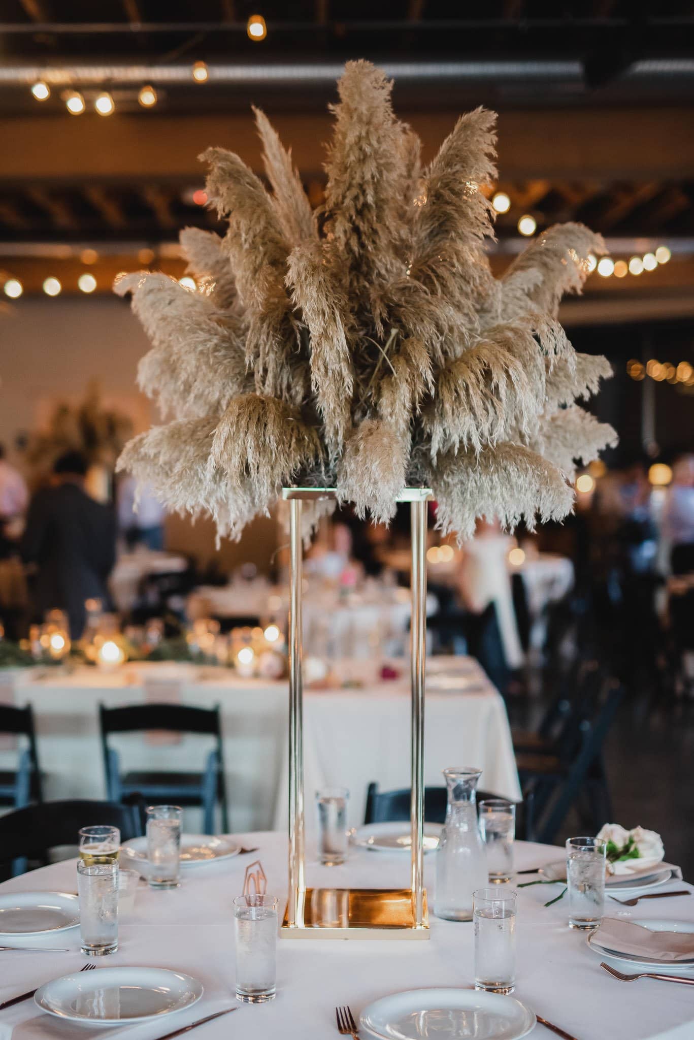 A tall centerpiece with pampas grass graces the dining table, surrounded by glasses and plates, creating a perfect setting for wedding photos at Castaway Portland. Warm lighting in the background adds to the inviting ambiance.