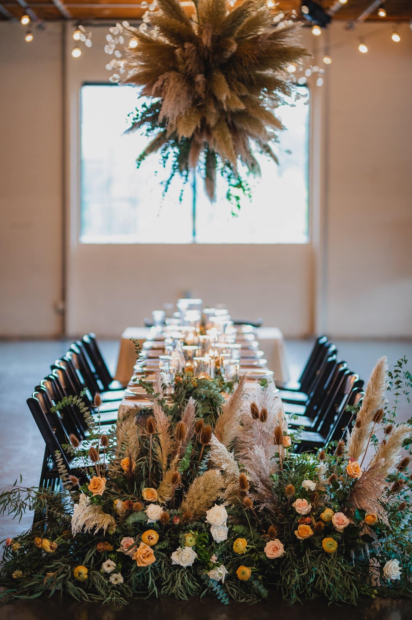 An elegant banquet table, adorned with floral centerpieces and hanging pampas grass, sits under a softly lit industrial ceiling. Perfect for capturing stunning wedding photos at Castaway Portland.