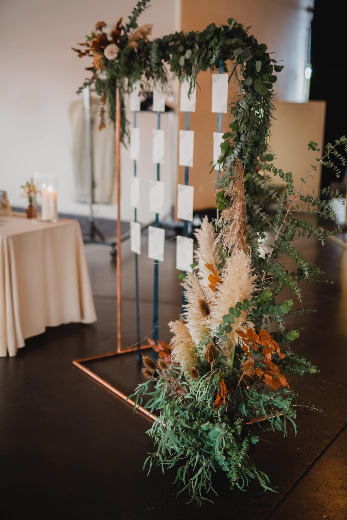 A wedding seating chart stand, embellished with greenery and pampas grass, is positioned near a draped table with candles, creating a picturesque scene perfect for wedding photos at Castaway Portland.