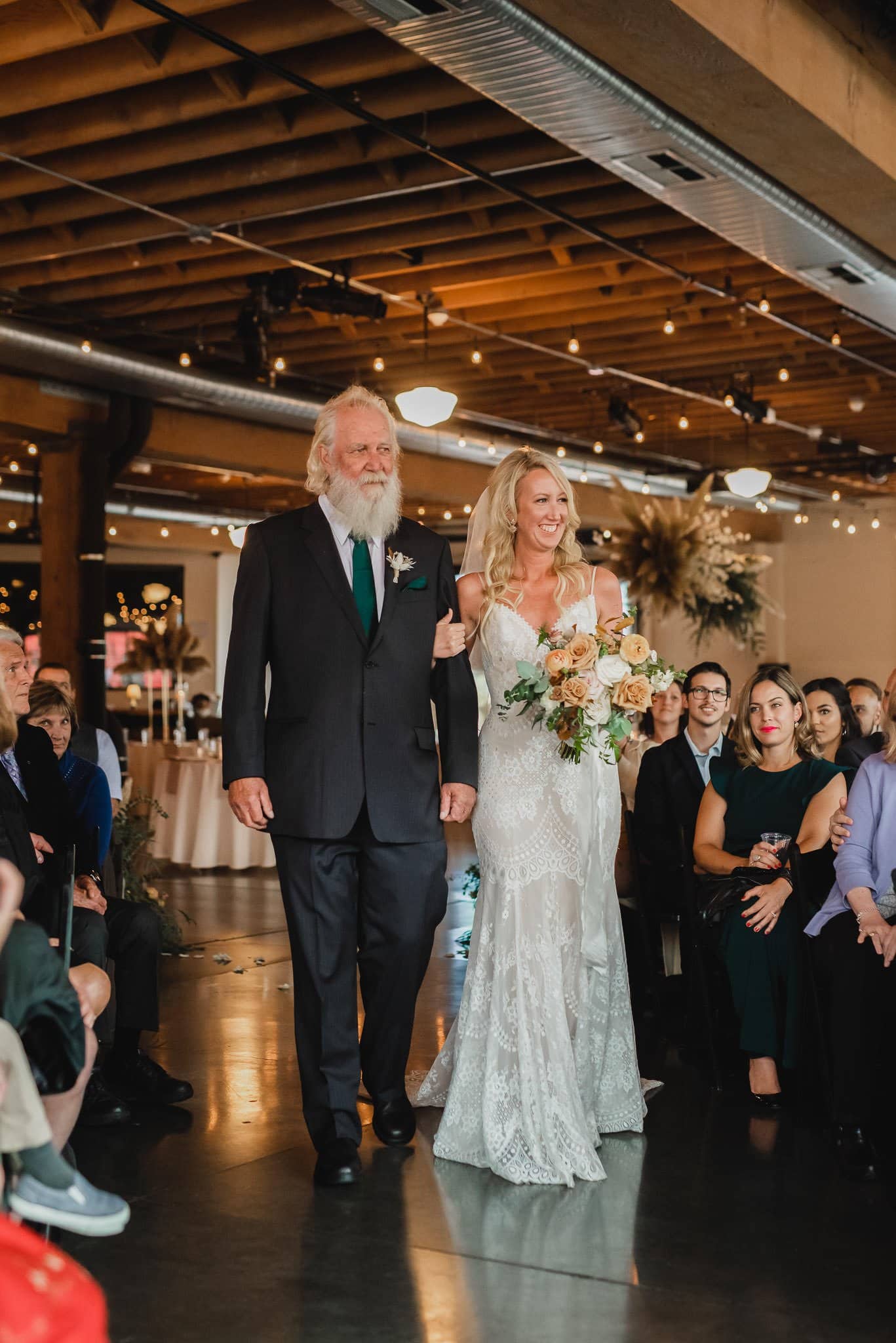 A bride in a lace dress walks down the aisle with an older man, carrying a bouquet. Capturing the moment, guests eagerly take wedding photos at Castaway Portland, seated on either side.
