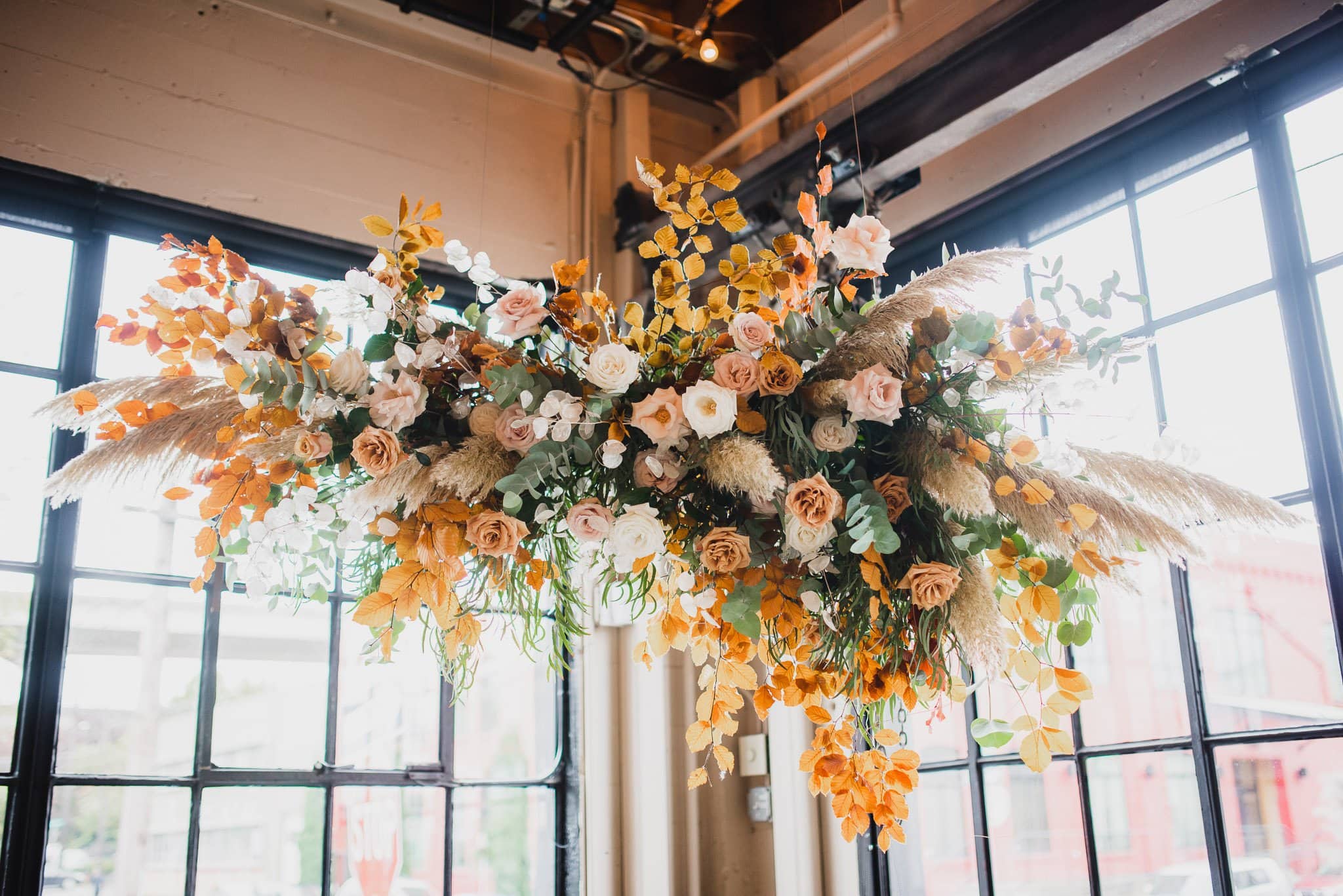 The floral installation, featuring orange leaves, white and pink roses, and pampas grass against large windows, creates a stunning backdrop for wedding photos at Castaway Portland.