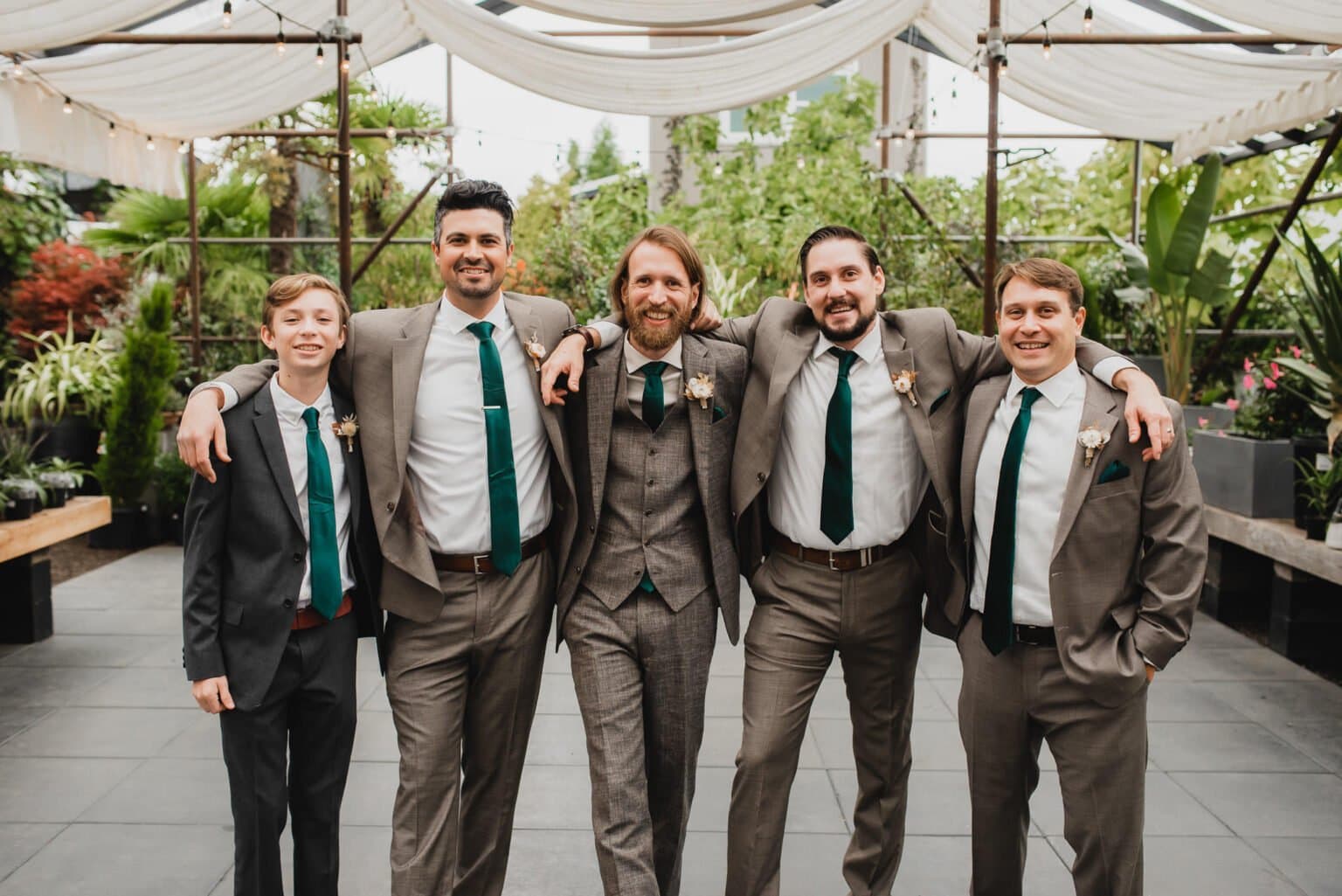 Five men in suits and ties smile with arms around each other in an outdoor setting filled with greenery and string lights, taking portraits at Blockhouse before a wedding at Castaway Portland.