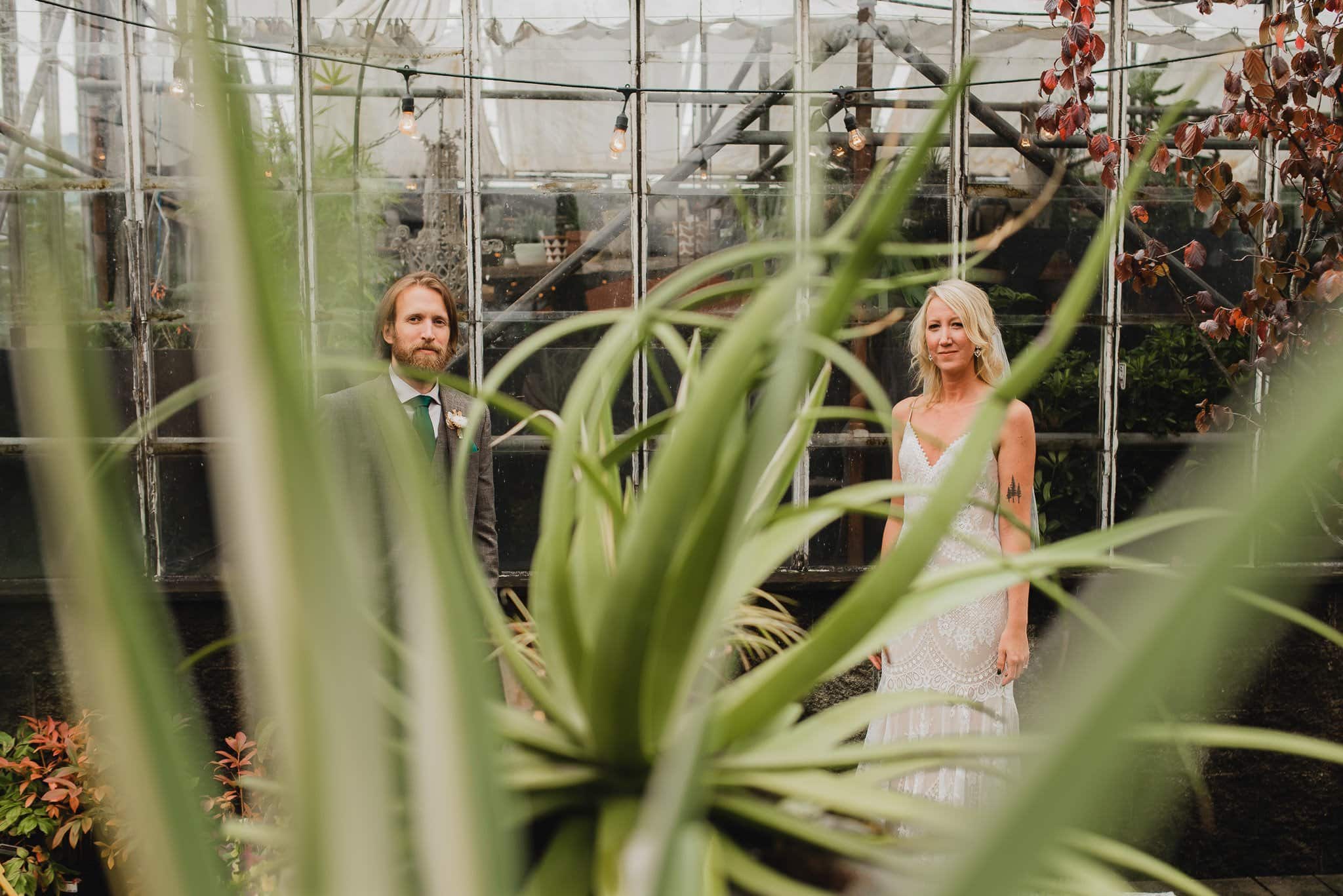 A couple poses in a greenhouse at Pomerius Nursery, partially obscured by the lush plant leaves in the foreground, just before their wedding at Castaway Portland.