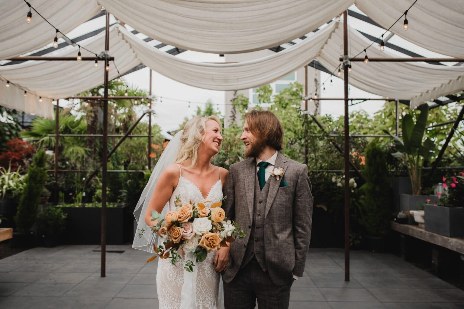 The bride and groom share a joyful smile under a draped canopy, holding a bouquet of roses. Their happiness is captured in beautiful portraits taken at Blockhouse before their wedding at Castaway Portland.