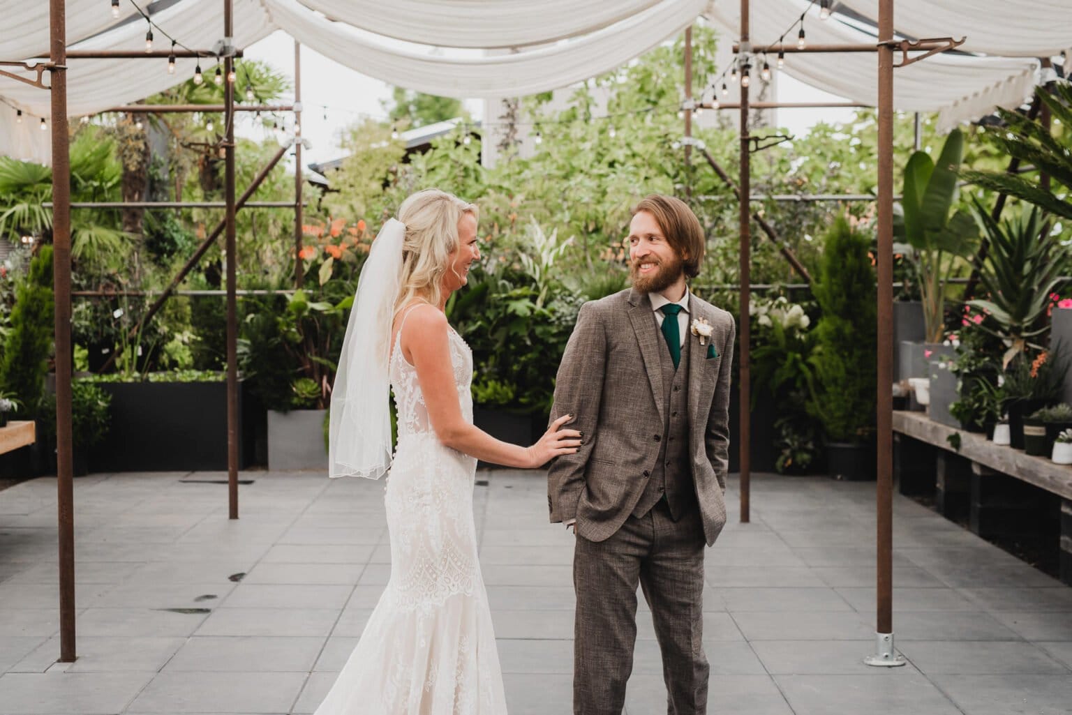 The bride in a white dress and veil smiles at the groom in a brown suit and green tie, standing under a canopy in a garden setting. They look forward to portraits at Blockhouse before their wedding at Castaway Portland.