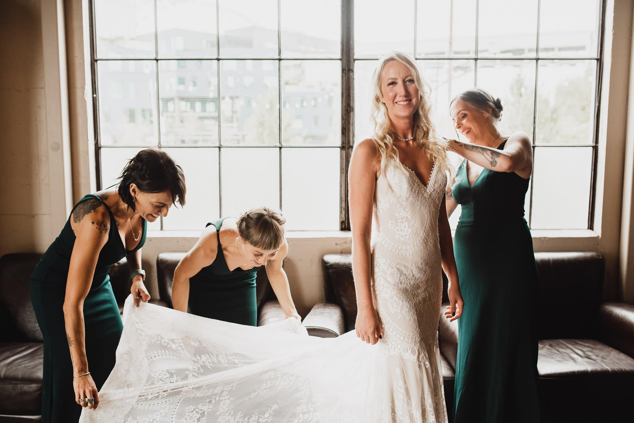 Bride in a lace dress smiles as three bridesmaids adjust her gown in front of large windows, capturing perfect wedding photos at Castaway Portland.