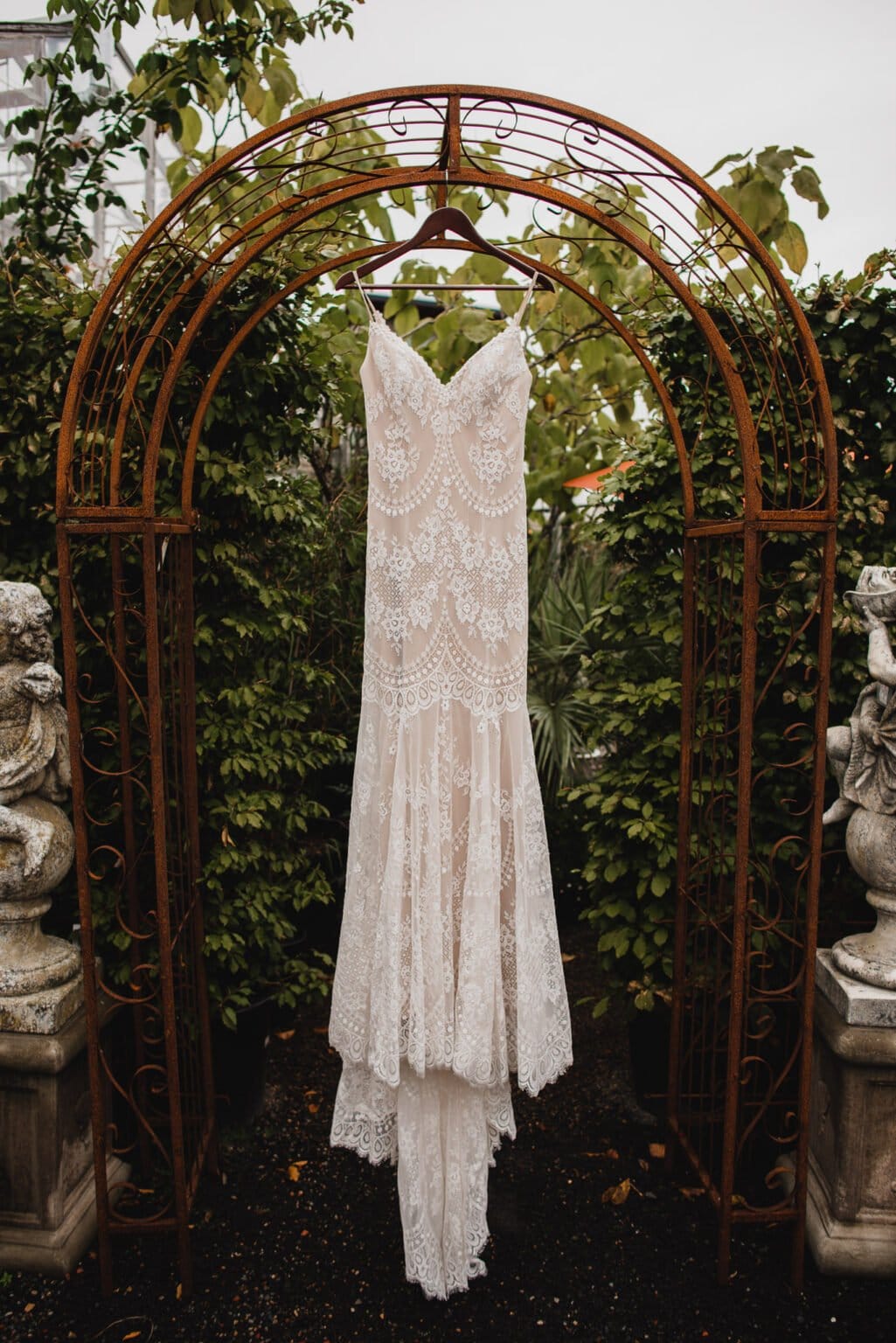 A lace wedding dress hangs on a rustic metal archway surrounded by greenery, capturing the perfect moment for wedding photos at Castaway Portland.