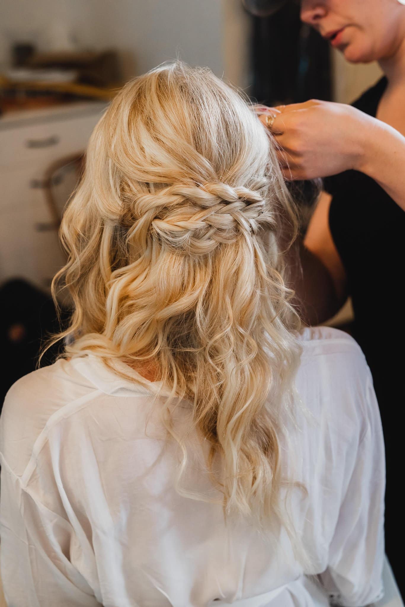 A blonde-haired bride with a braided half-up hairstyle is getting ready at a nearby salon during a Castaway wedding, assisted by a skilled hairstylist.