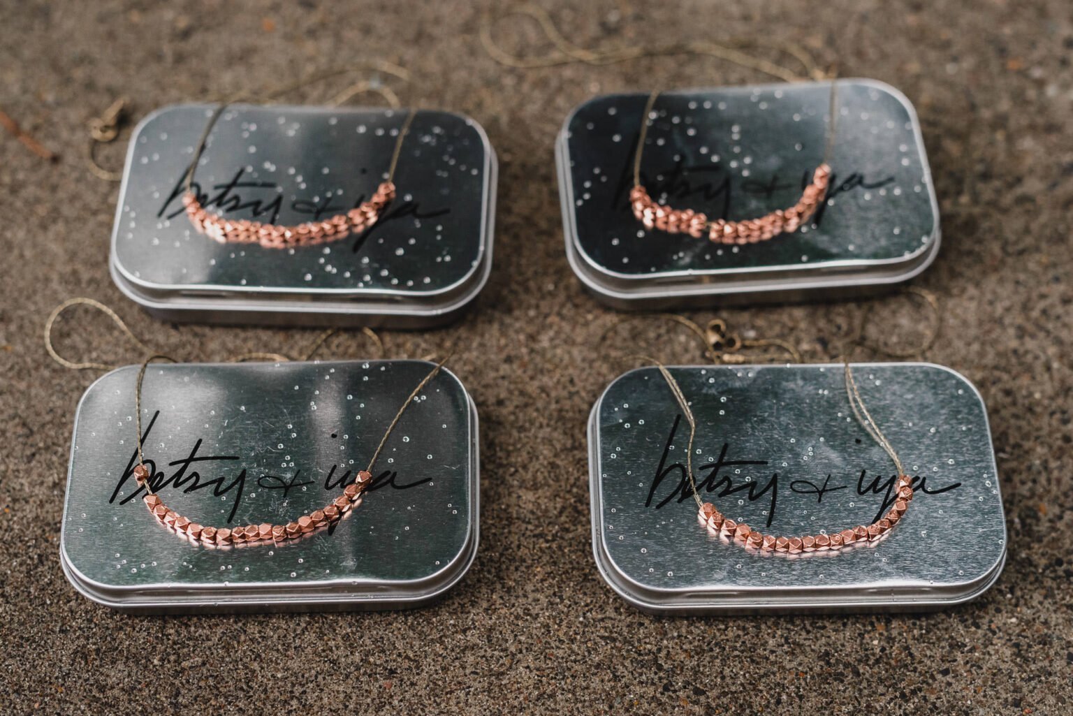 Four metallic tins on a textured surface, each showcasing a copper beaded necklace. Perfect for getting ready at a nearby salon during a Castaway wedding.