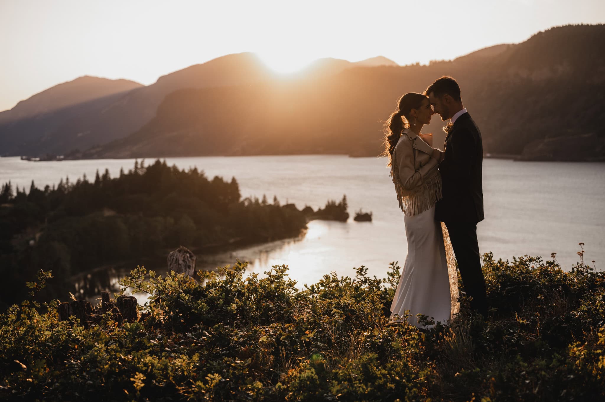 A couple embracing at sunset by a lake, surrounded by hills and foliage against a golden sky, creates a perfect setting for wedding photos at The Griffin House.
