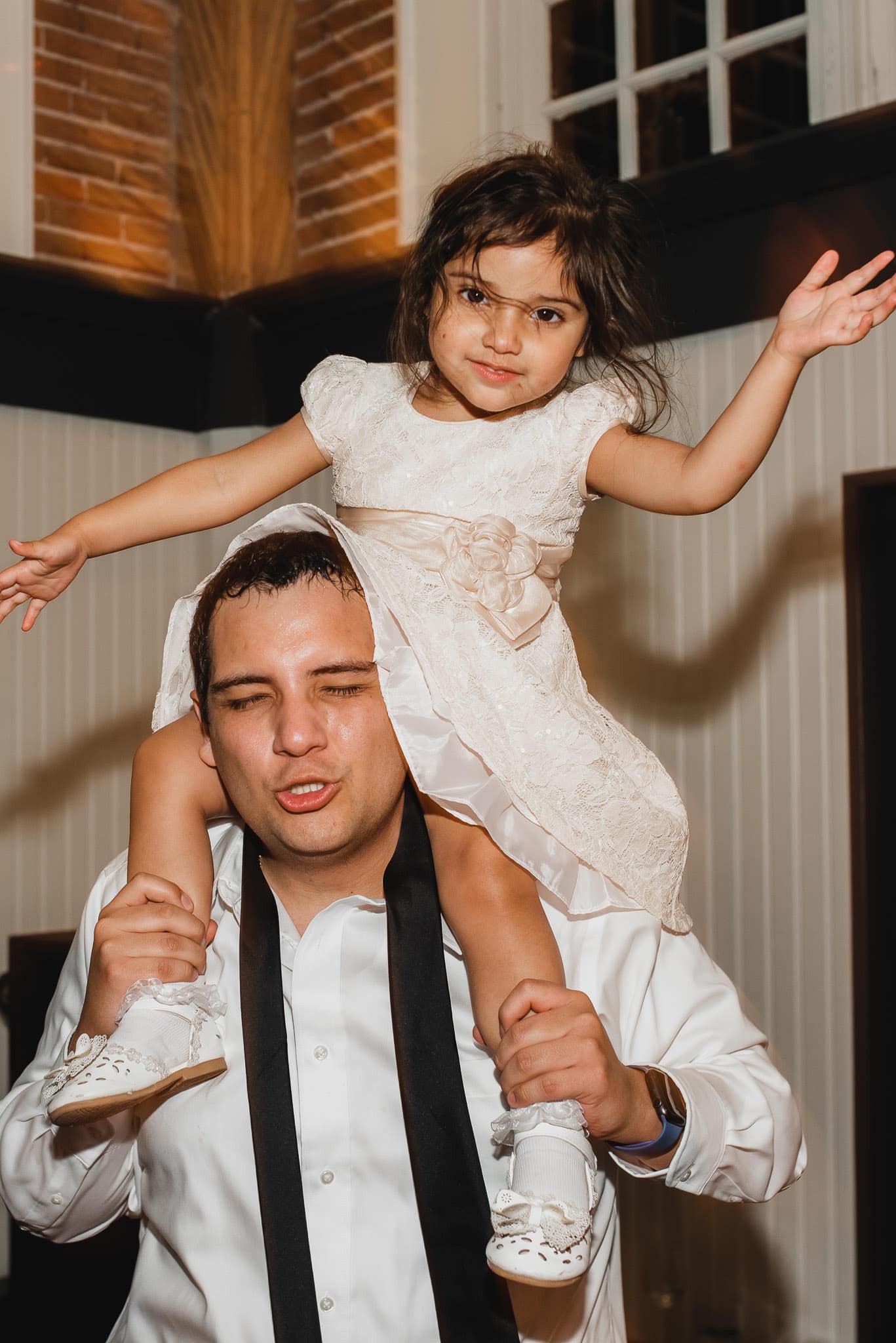 A man joyfully smiles with a little girl in a white dress perched on his shoulders, surrounded by the warm glow of the room. Its as if theyre celebrating a wedding at the Evergreen, their laughter and happiness filling the space.