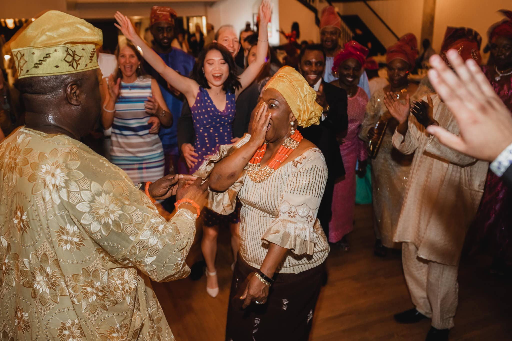 At a vibrant wedding at the Evergreen, a joyful group of people in colorful attire dance and celebrate, with one woman in front reacting enthusiastically.