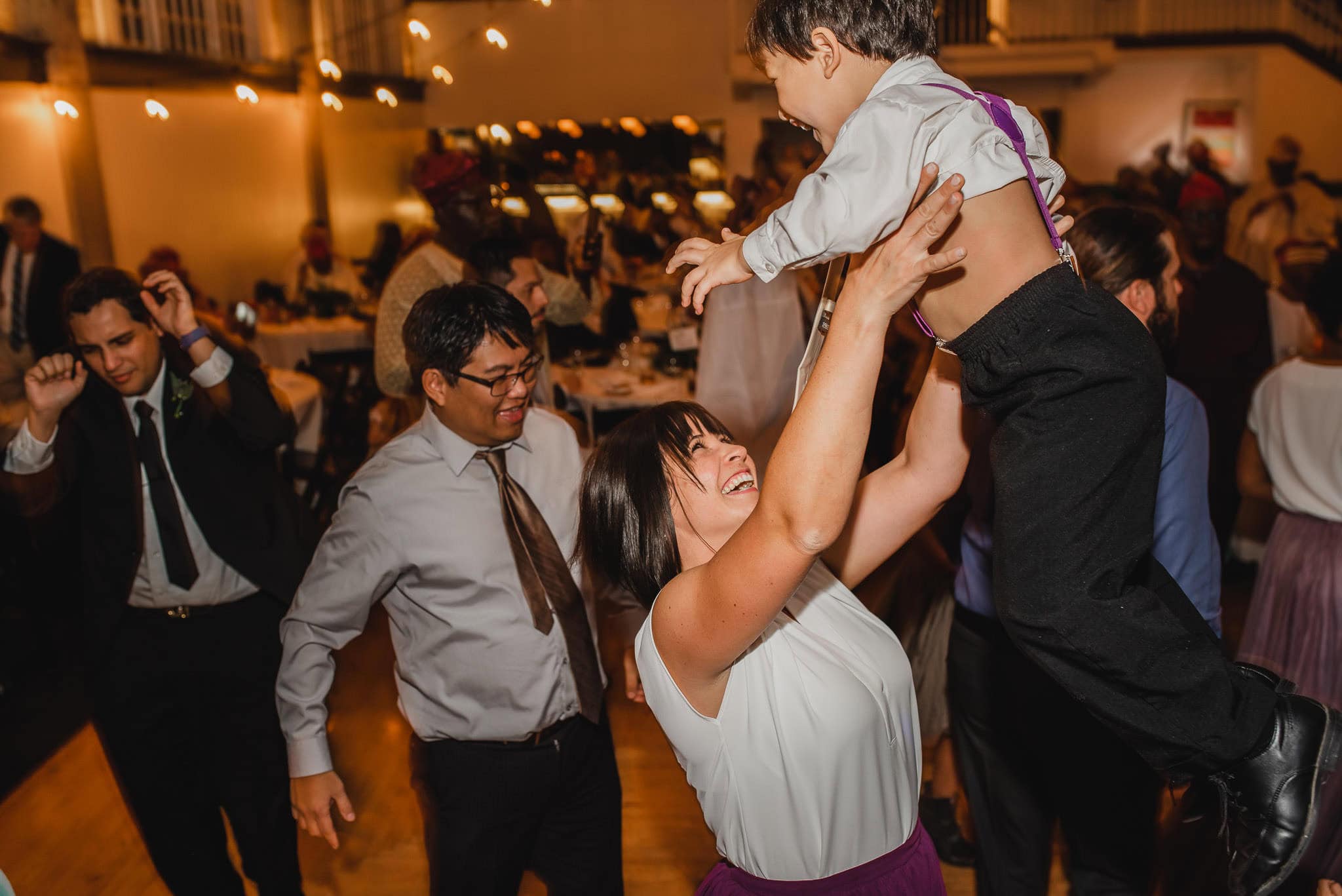 At a wedding at the Evergreen, a woman joyfully lifts a child amid an indoor celebration, surrounded by dancing people.