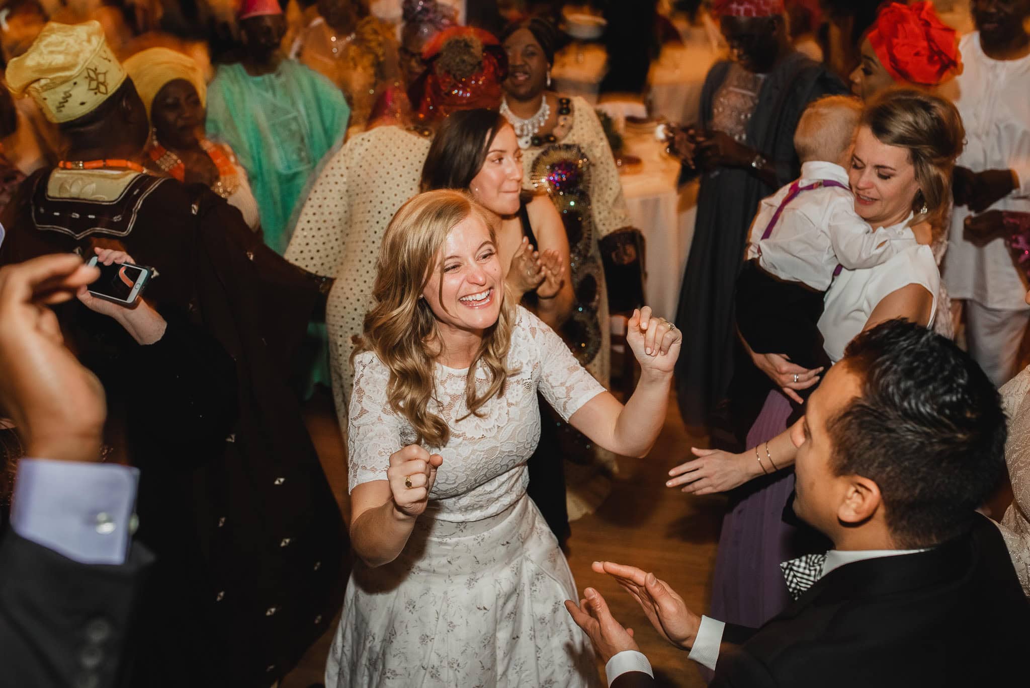 At a lively indoor event, a woman in a white dress dances joyfully among guests at the wedding at the Evergreen.