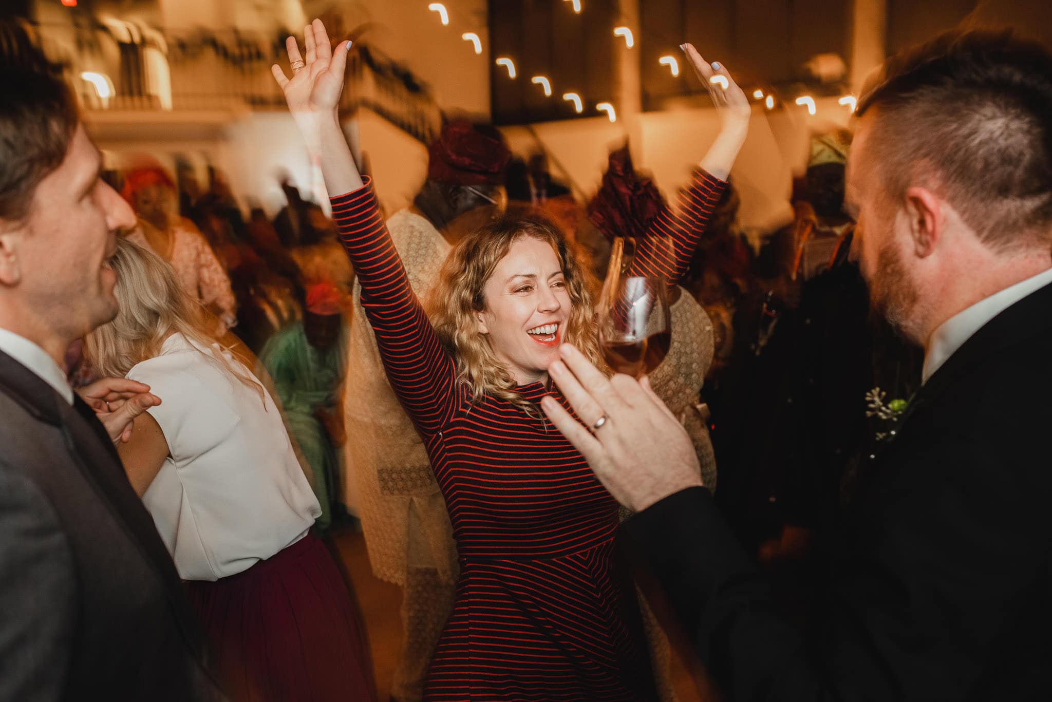 A woman in a striped dress dances joyfully with arms raised, surrounded by guests at a lively wedding at the Evergreen.
