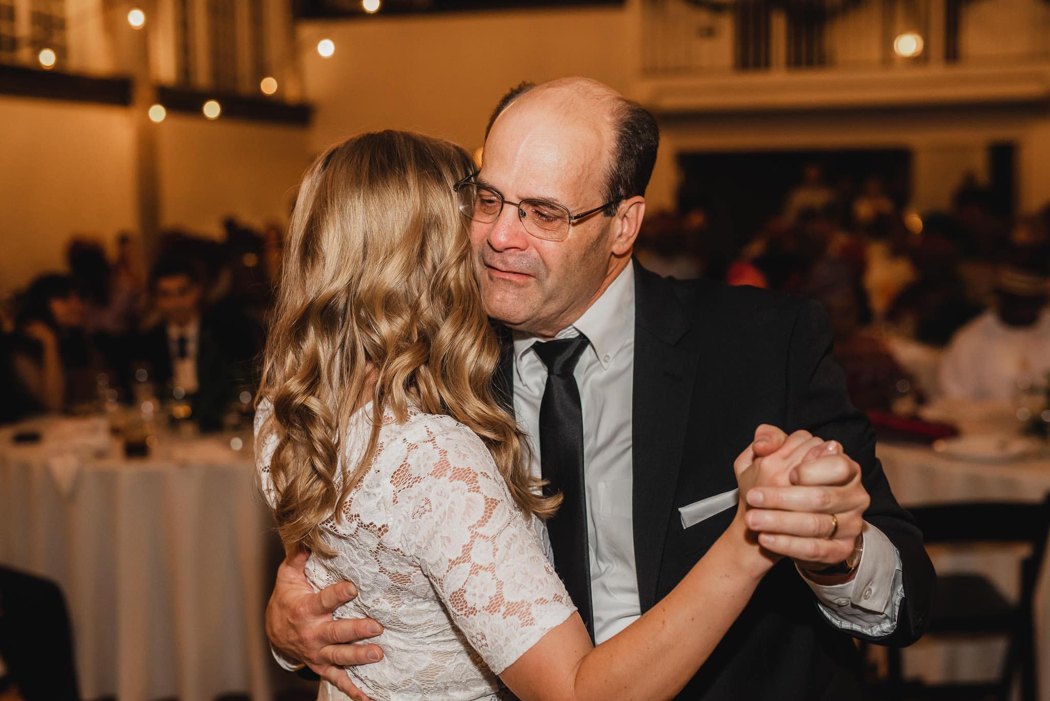 At the Evergreen, a man in a suit lovingly dances with a woman in a white lace dress at an indoor event, creating a magical moment together.