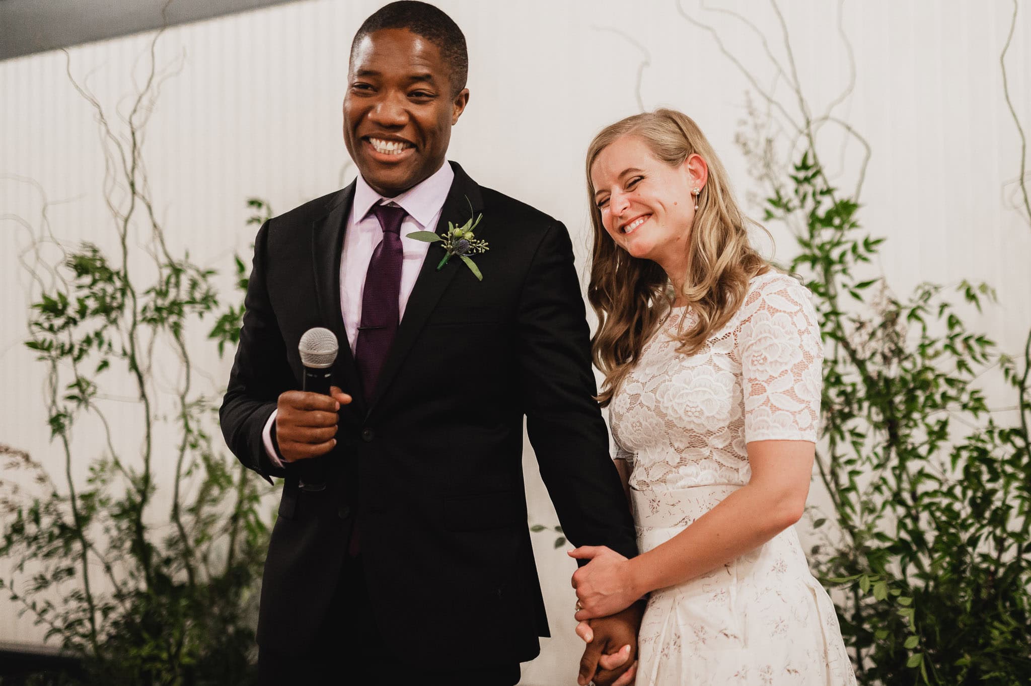 A smiling couple stands holding hands at the wedding at the Evergreen; the man holds a microphone. They are surrounded by lush greenery.