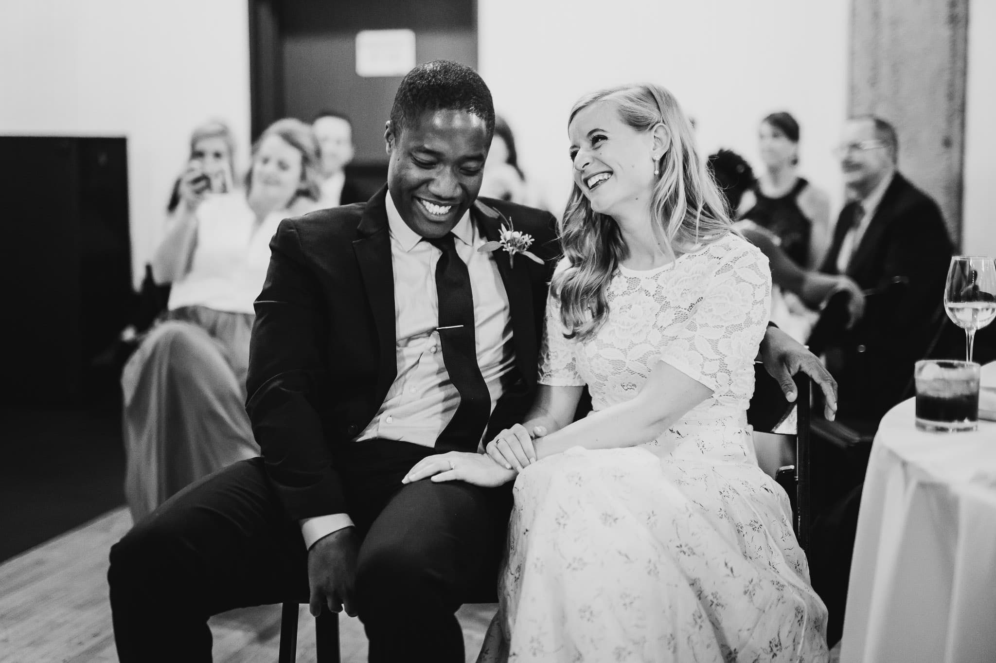 A joyful couple sitting close and smiling, surrounded by people in a black-and-white photo, capturing a special moment at the wedding at the Evergreen.