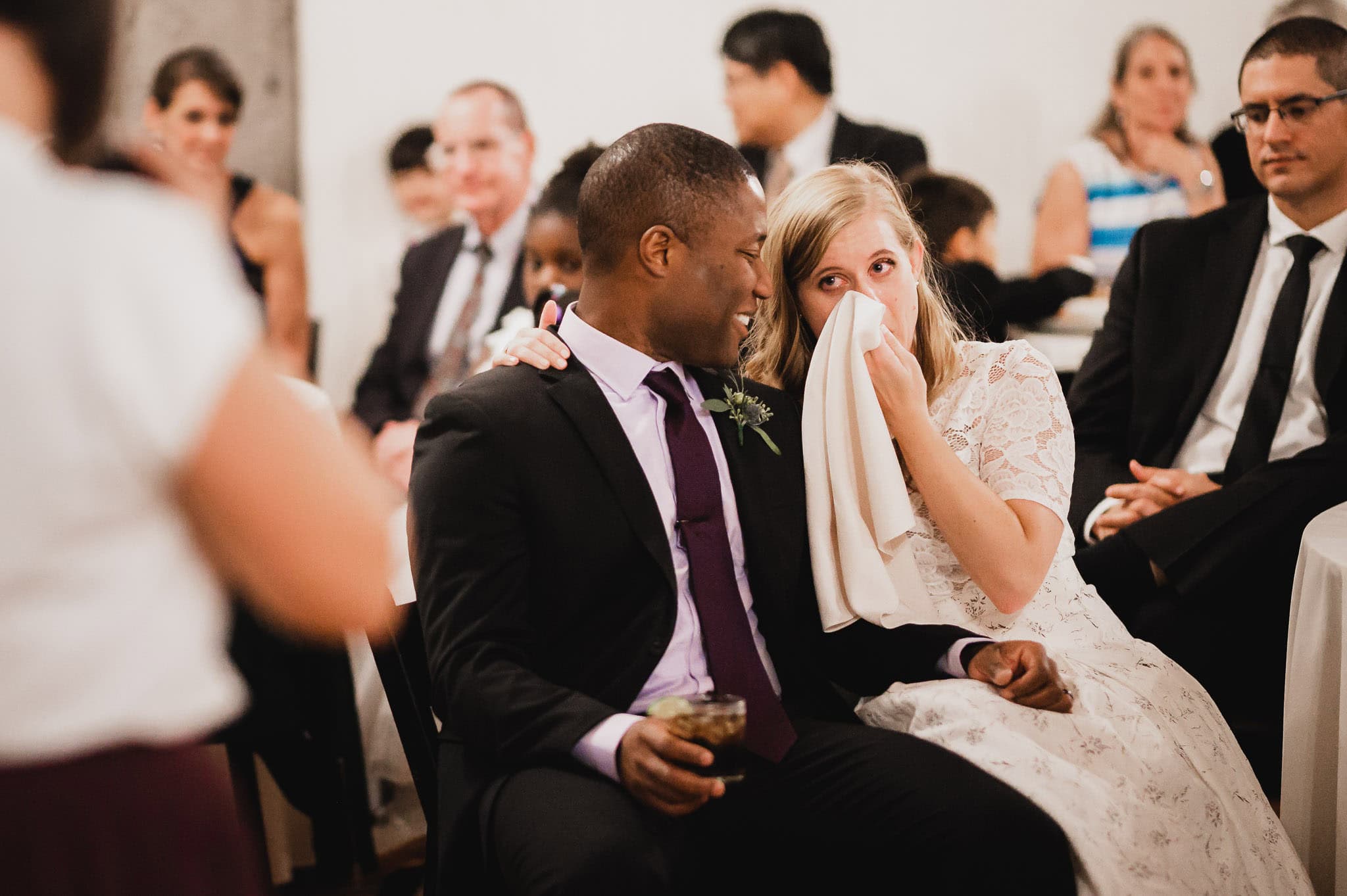 At the wedding at the Evergreen, a couple sits closely; the woman wipes her tears with a napkin while guests watch with smiles.