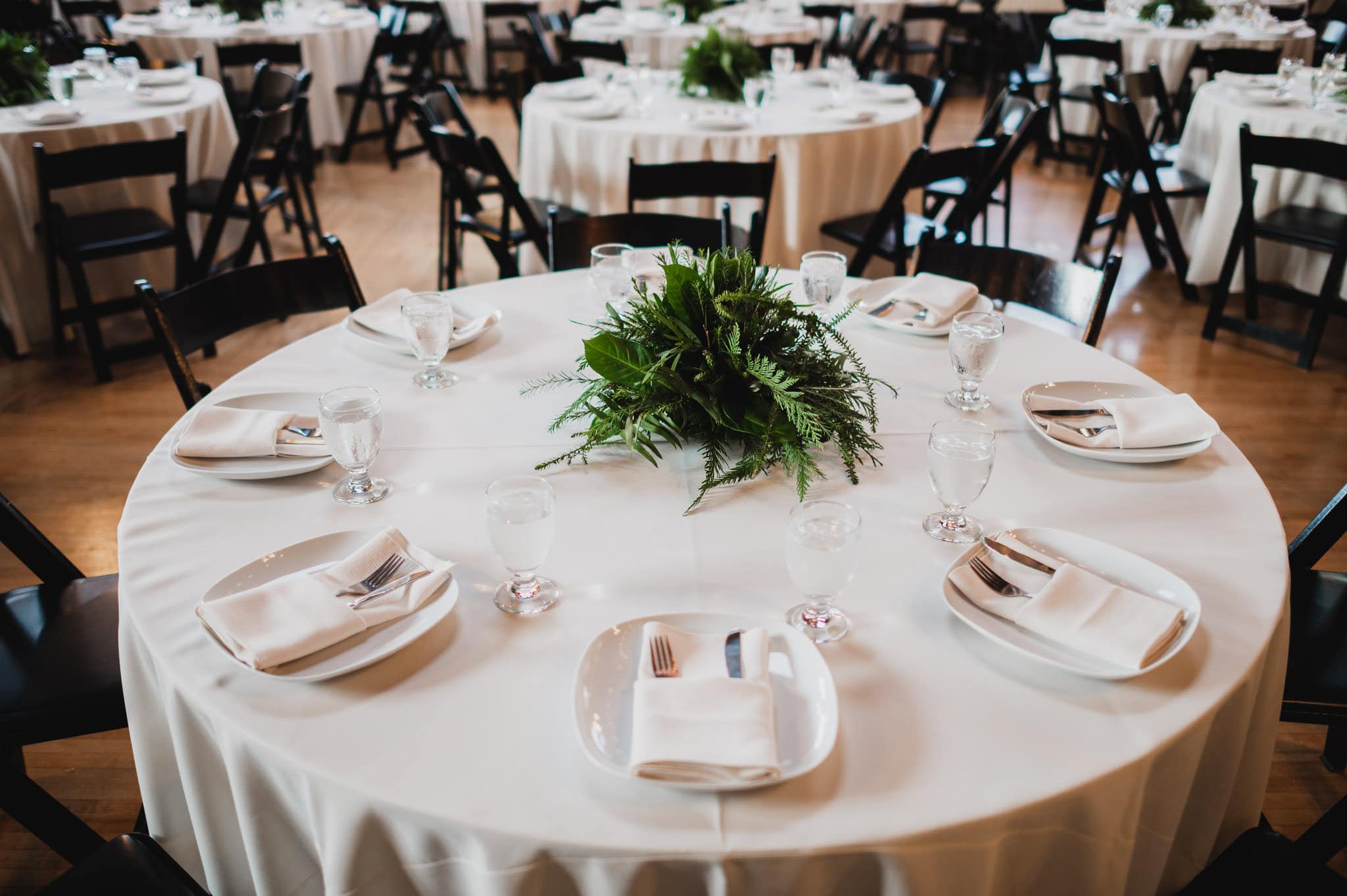 The round table set for a wedding at the Evergreen features a white tablecloth, plates, cutlery, glasses, napkins, and a green fern centerpiece.