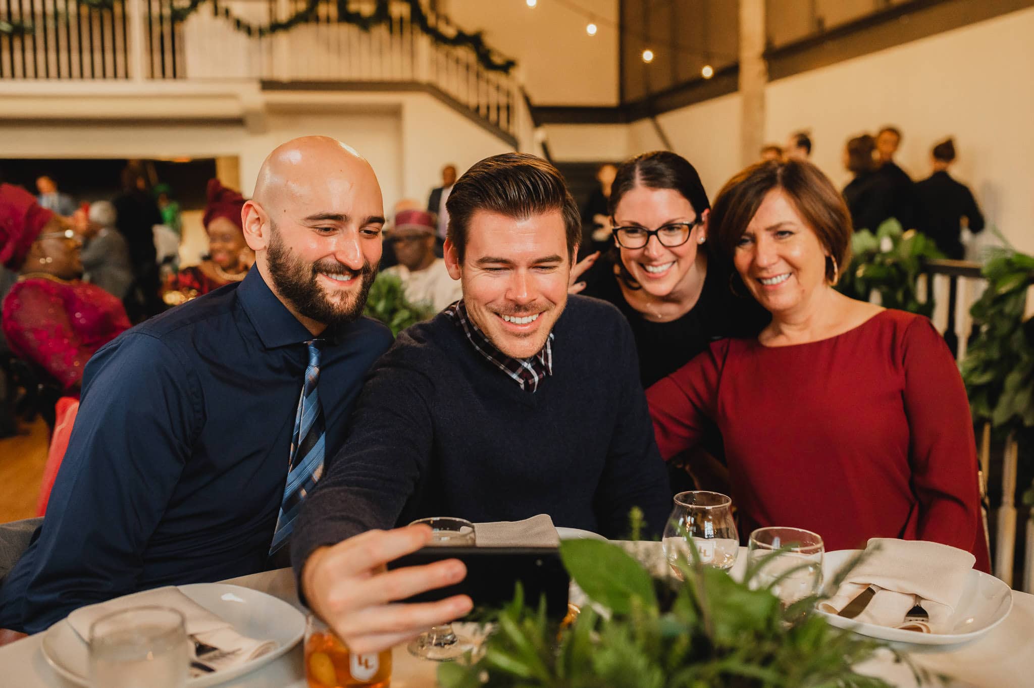 Four people sitting at a dining table, smiling and taking a selfie at an indoor event with festive decor, celebrating a wedding at the Evergreen.