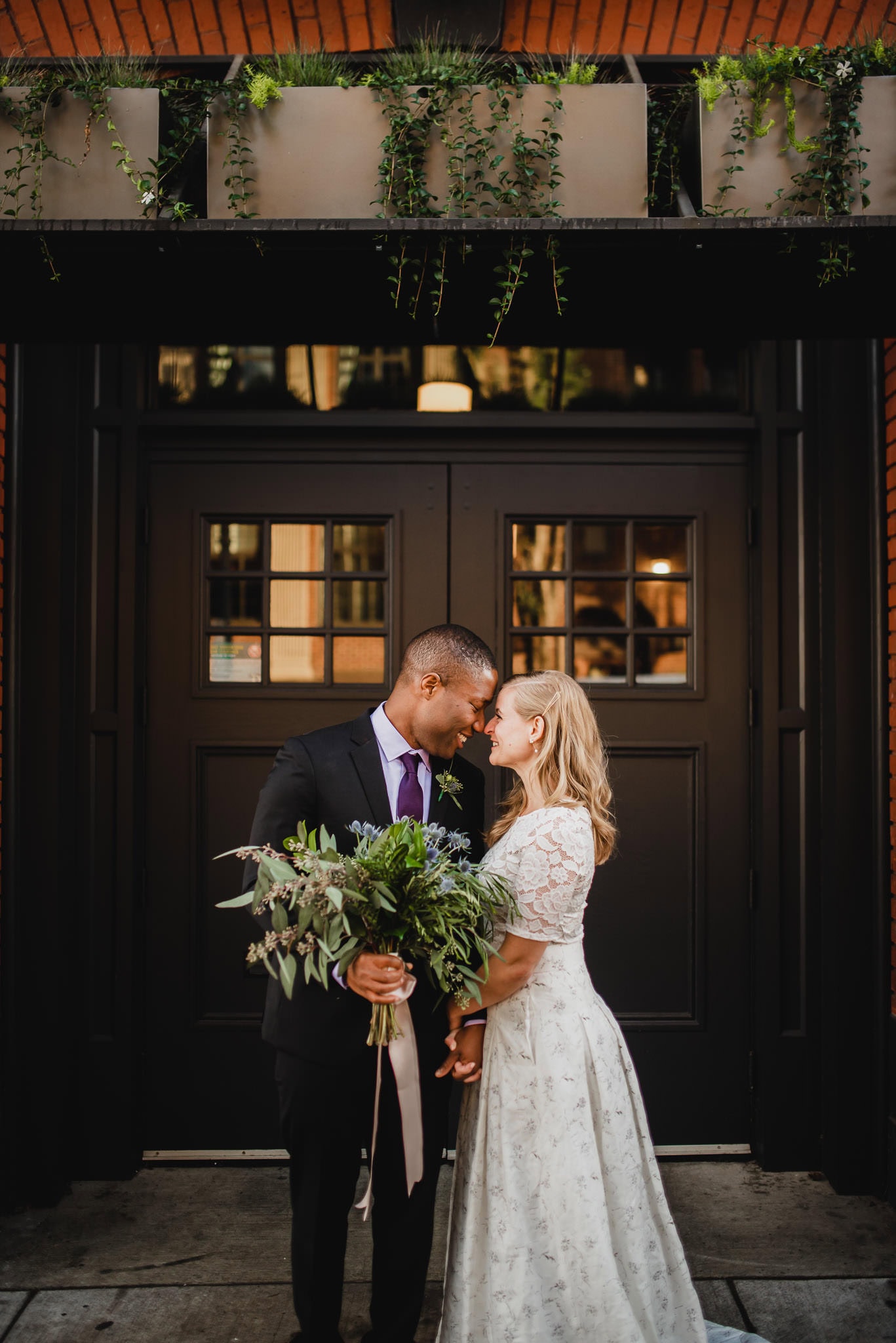 At the wedding at the Evergreen, the bride and groom lovingly lean their heads together, holding a bouquet in front of large black doors adorned with greenery.