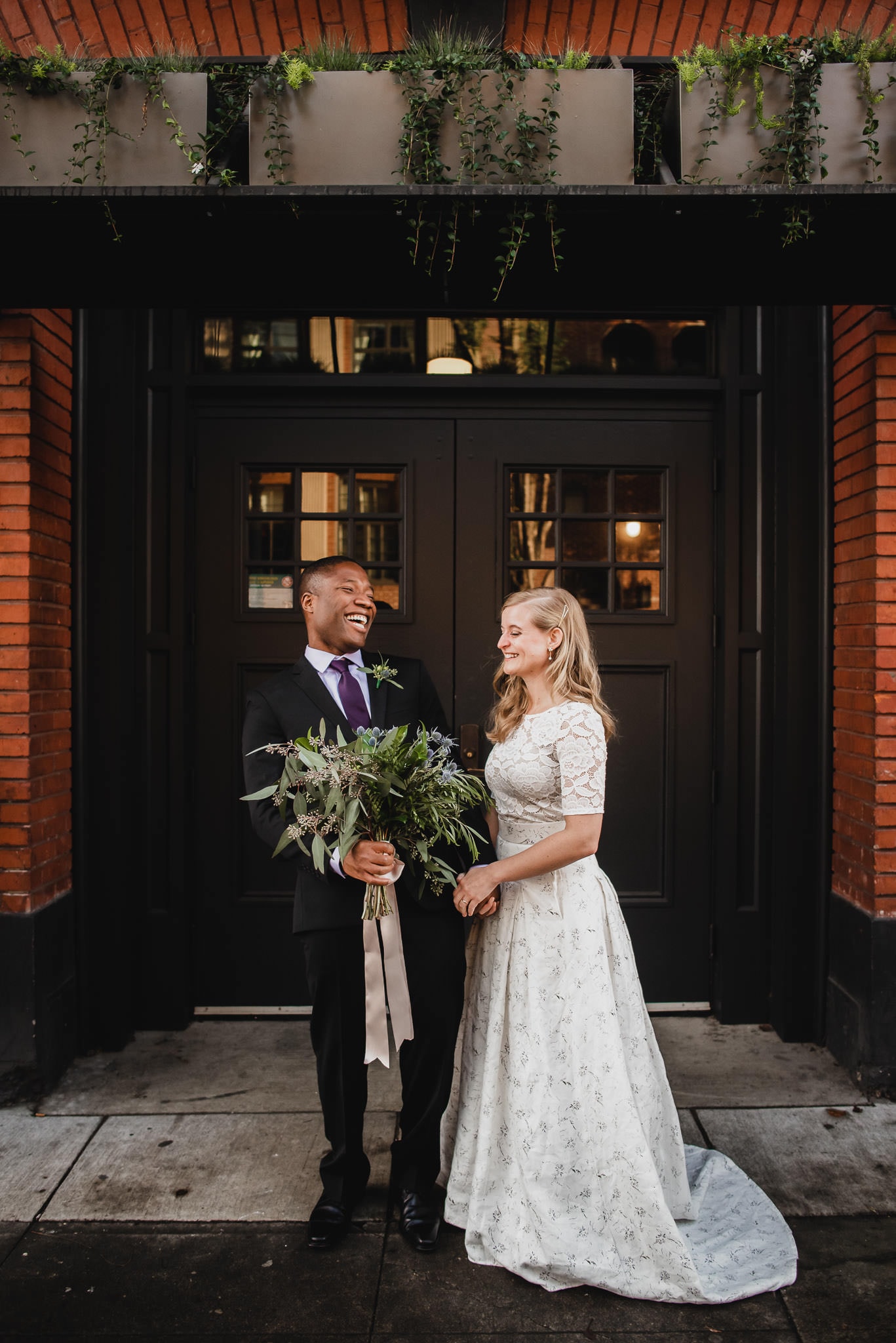The bride and groom stand smiling before a brick wall, holding a bouquet, capturing the joy of their wedding at the Evergreen.