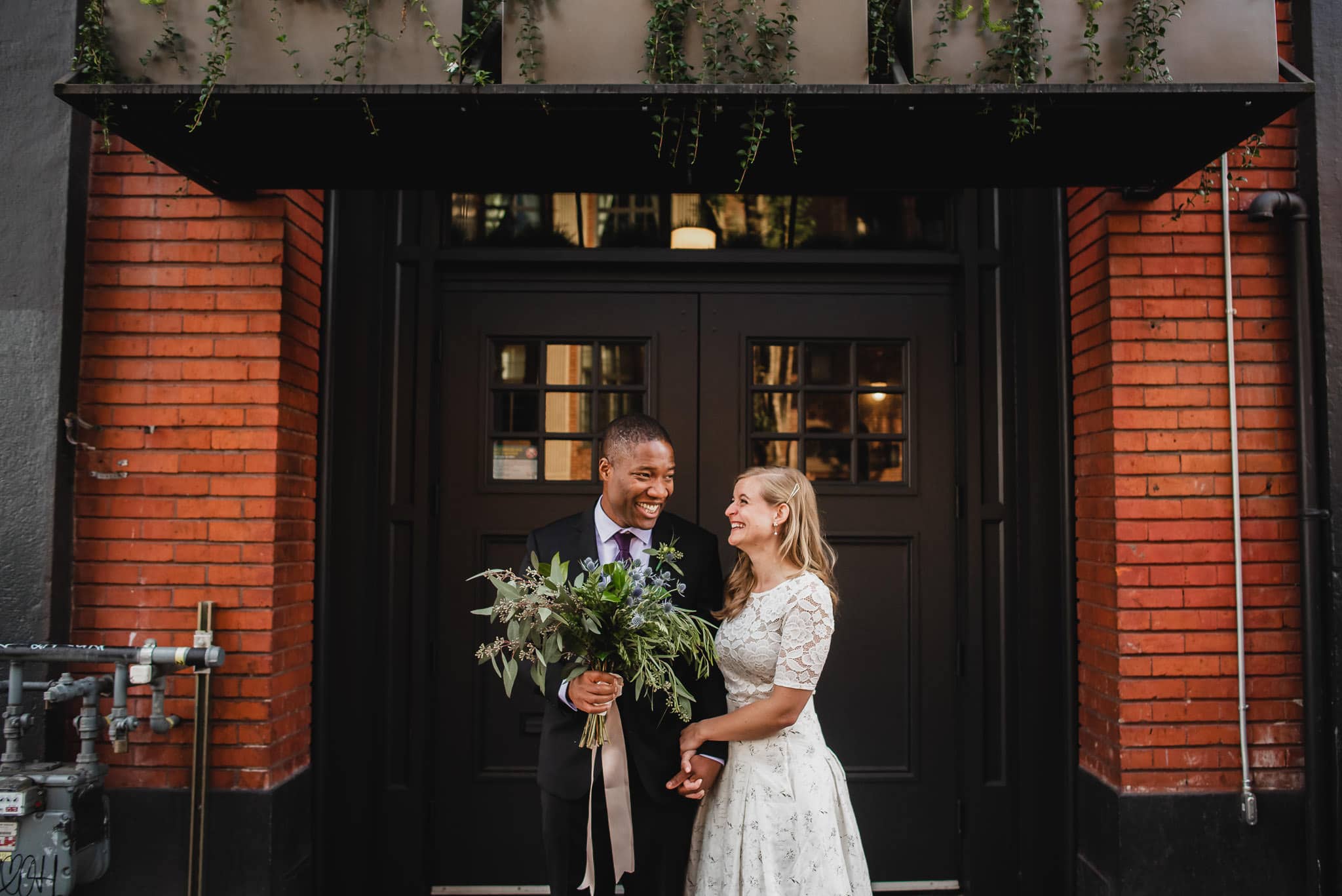 The couple, dressed in wedding attire, smiles joyfully in front of a brick building adorned with greenery at the Evergreen.