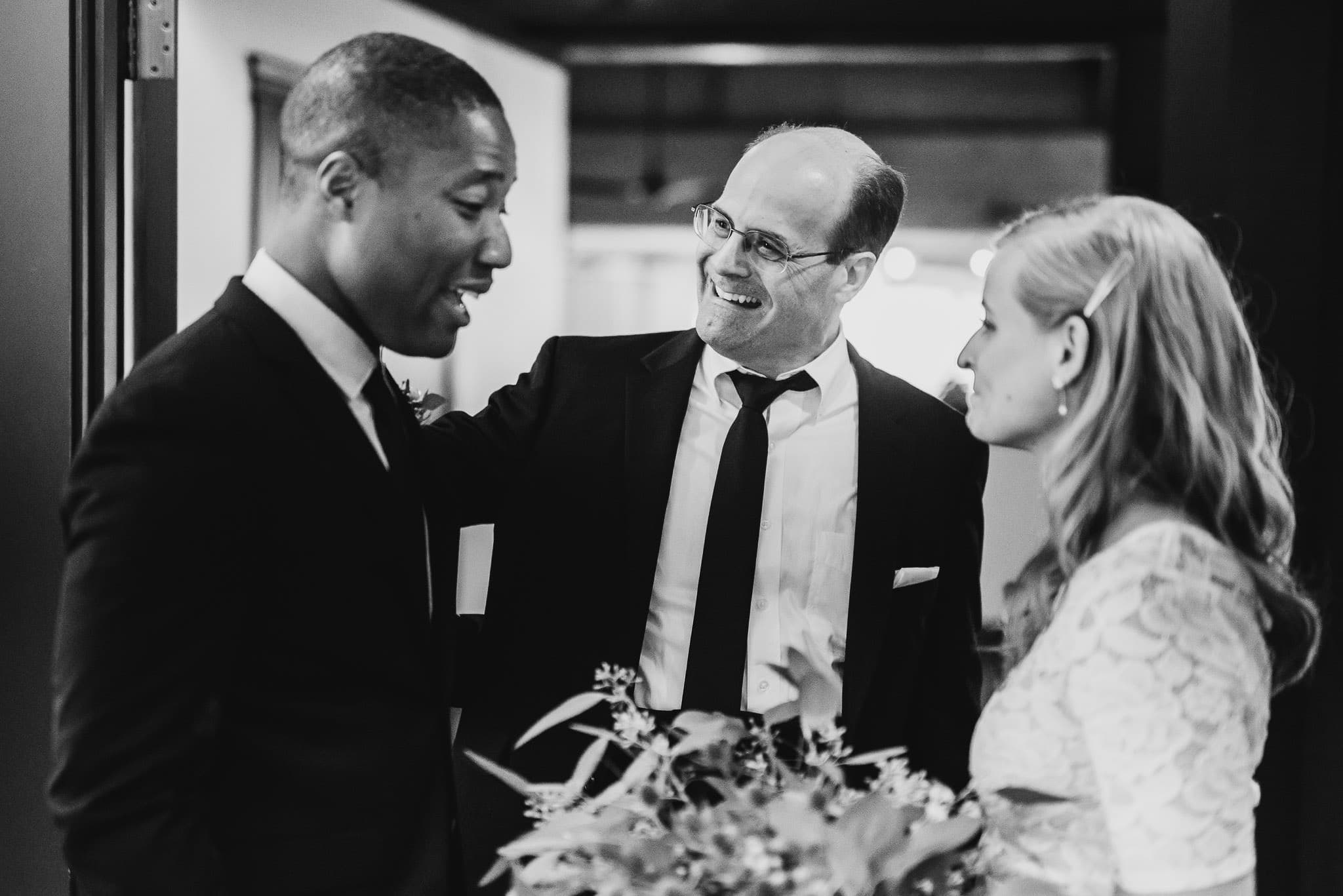 Three people in formal attire share a conversation indoors at the wedding at the Evergreen, with one holding a bouquet.