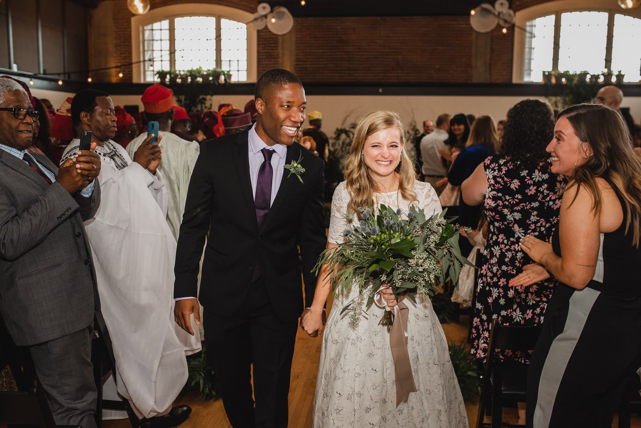 The bride and groom are all smiles as they hold hands and walk down the aisle at their wedding at the Evergreen, surrounded by applauding guests.